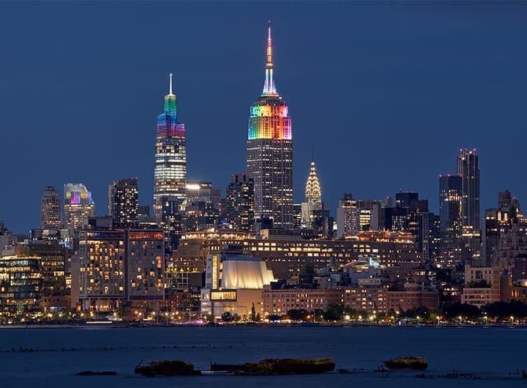 Die berühmte New Yorker Skyline bei Nacht, einige Hochhäuser und Wahrzeichen leuchten in Regenbogenfarben für die NYC Pride